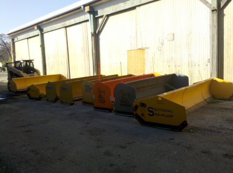 2003 Ford F-250 - A collection of snow plows are lined up in front of a large building, with a skid steer loader visible on the left.