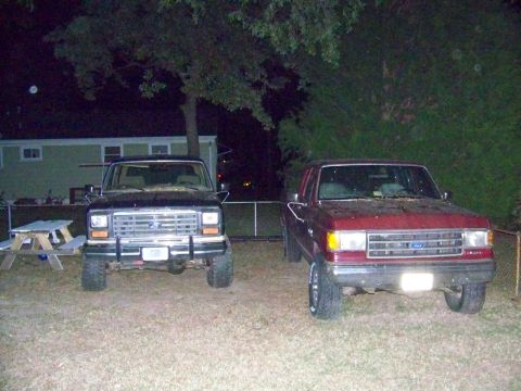 1985 Ford Bronco - Two Ford Broncos are parked side-by-side in a yard at night, with a house and trees in the background.