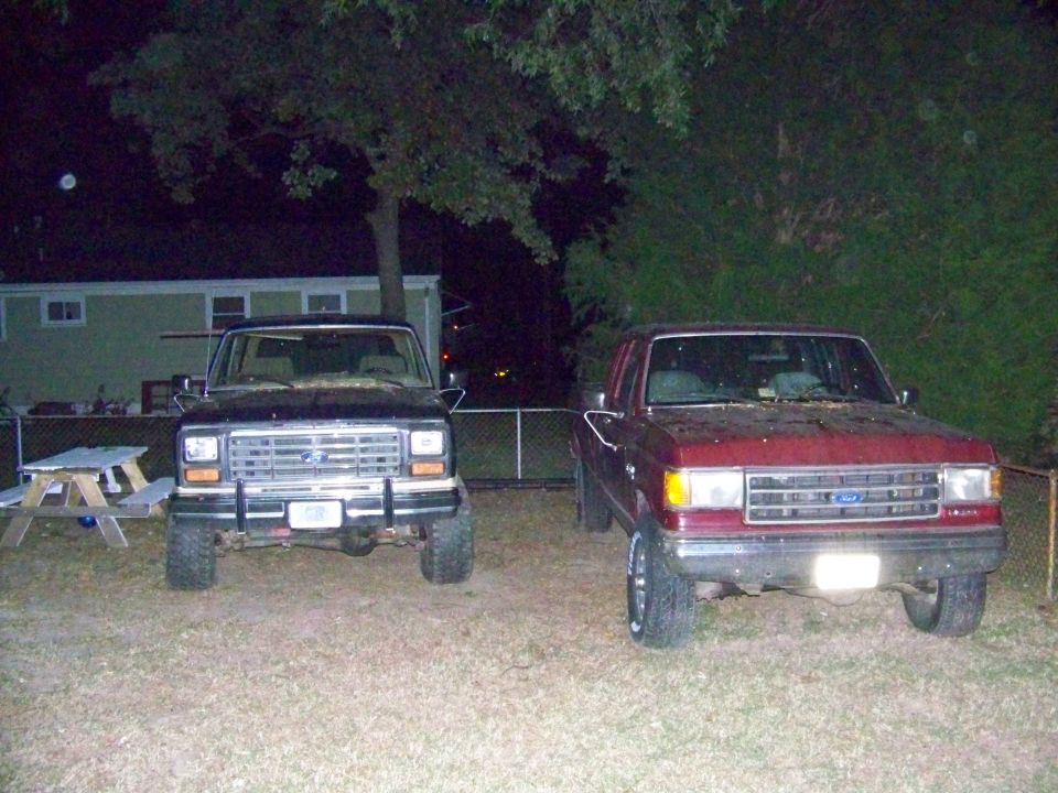 Two Ford Broncos parked in a yard at night. The one on the left is black, and the one on the right is red. Both are facing forward