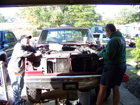 1985 Ford Bronco - Two men work on the engine of a red pickup truck, which has its hood and front grille removed.
