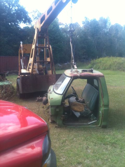 1993 Ford Bronco - A crane is lifting a green truck cab off its chassis in a grassy yard.