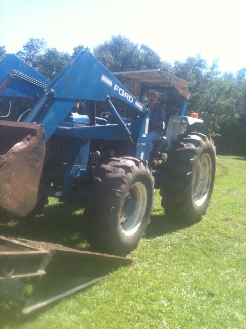 1993 Ford Bronco - A man drives a blue Ford tractor equipped with a front loader across a grassy area under a clear sky.