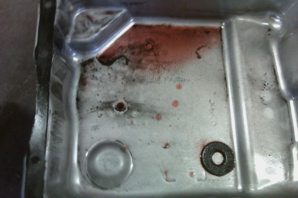 Inside of a metal transmission pan with red fluid residue, sludge, and a debris-covered round magnet