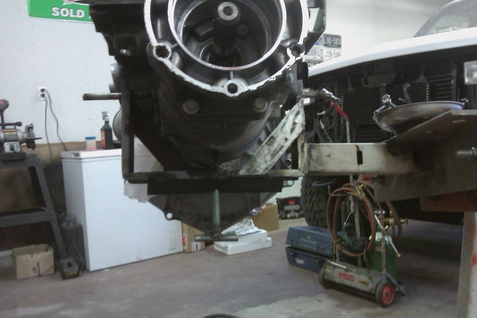 Removed automatic transmission on a stand in a garage, white vehicle front end visible at right