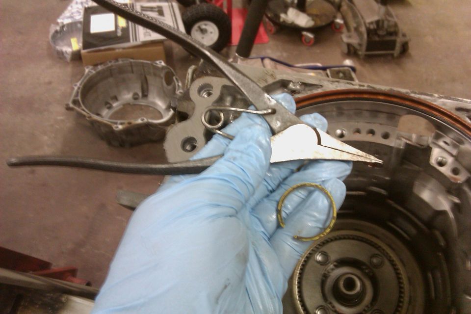 Gloved hand holding pliers and a snap ring over disassembled transmission components