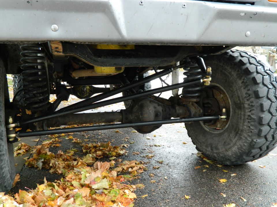 Undercarriage of a 1978 Ford Bronco showing front suspension, axle, and tire with fallen leaves on the ground