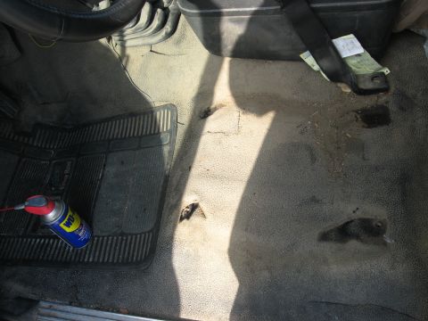 1981 Ford Bronco - The interior floor of a vehicle shows wear and tear, with a black rubber mat and a can of WD-40 visible.