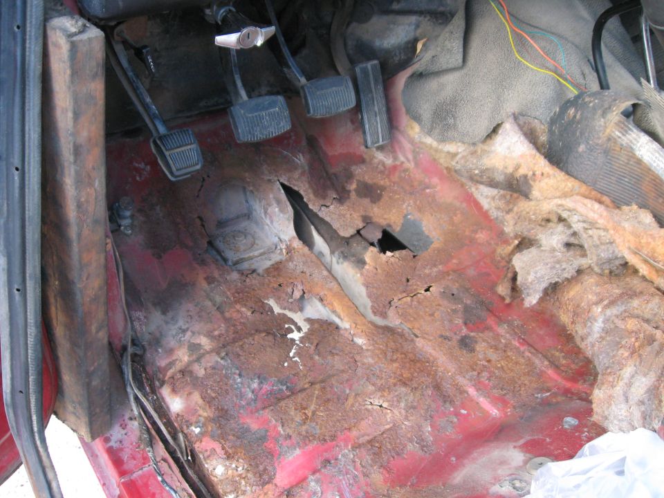 vehicle interior showing heavily rusted floor pan with holes, pedals visible