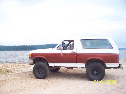 1989 Ford Bronco - A two-tone red and white Ford Bronco is parked on a gravel shore with a lake in the background.