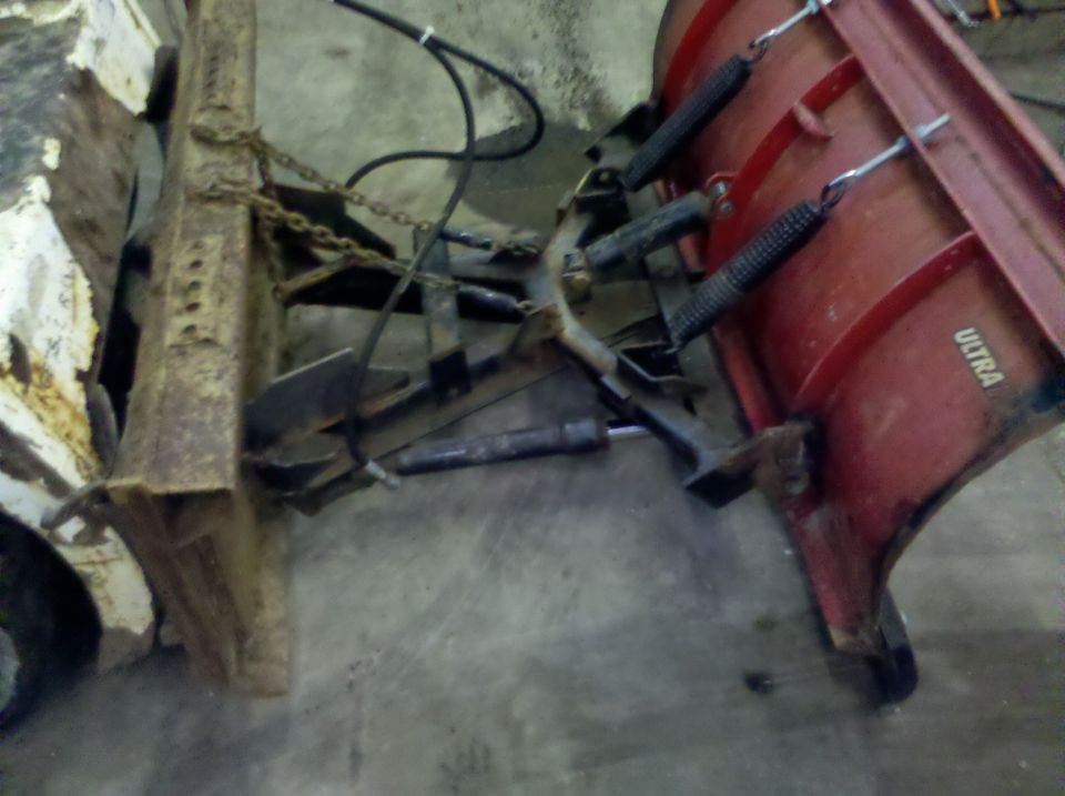 Red snow plow with "ULTRA" text attached to a rusty metal frame and chains