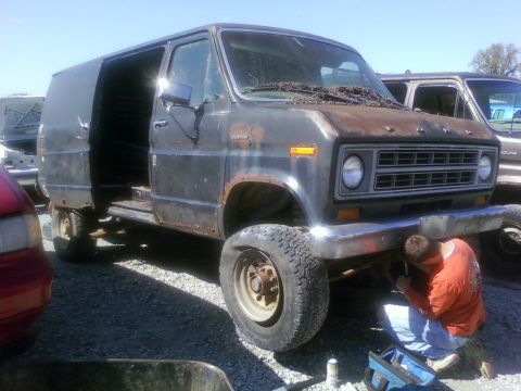 1989 Ford Bronco - A person works on the front bumper of a heavily rusted van parked outdoors.