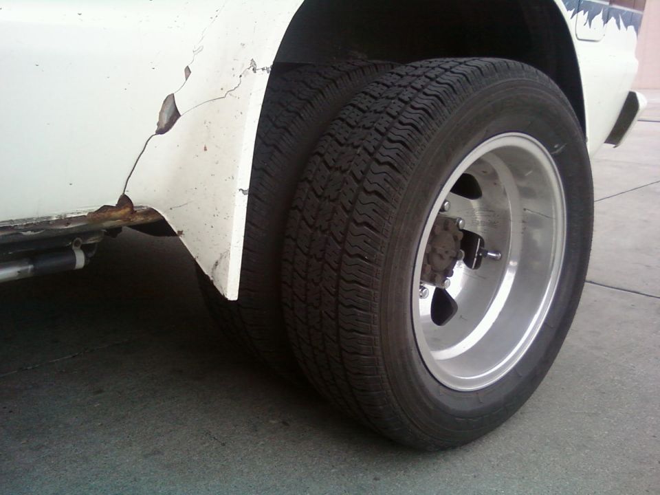 Close-up of dual rear wheels on a white vehicle with rust and peeling paint on the fender