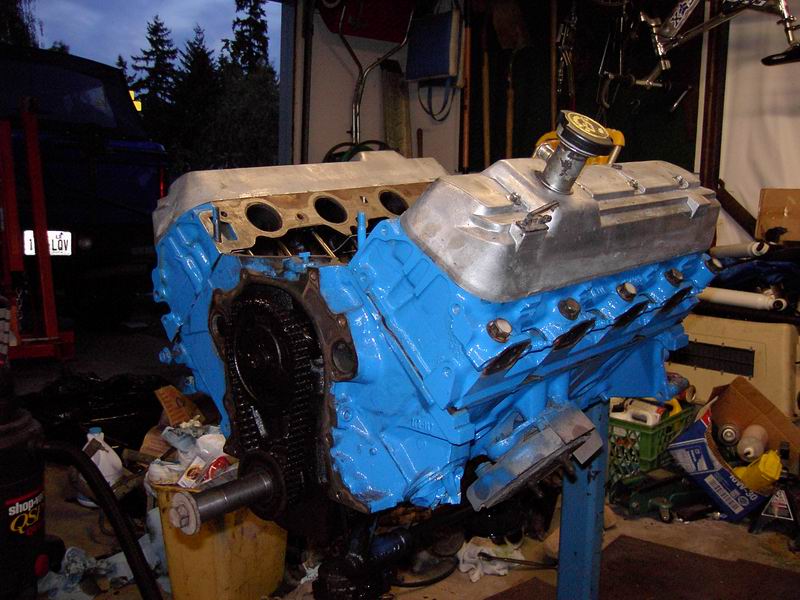 Blue-painted V8 engine block on a stand in a garage, one aluminum valve cover on, opposite head deck exposed.
