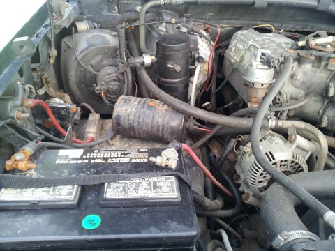 1995 Ford Bronco - The engine bay of a vehicle shows a battery, alternator, and numerous hoses and electrical components.