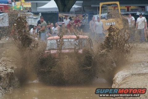 1978 Ford Bronco - A vehicle navigates a challenging mud pit, creating a massive spray as a tractor watches from the side.
