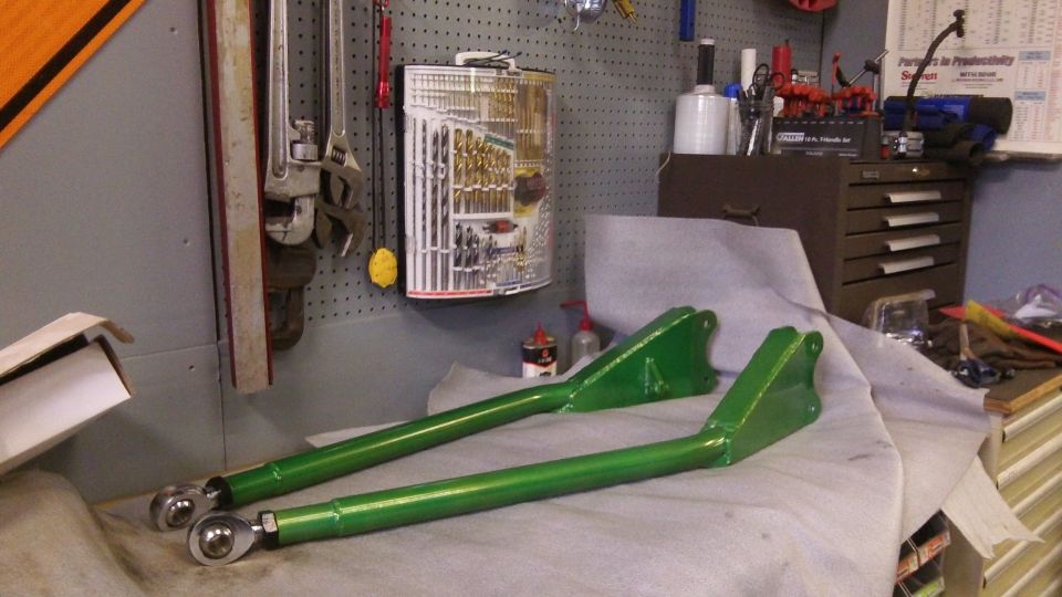 Pair of green metal suspension control arms with rod ends on a workbench