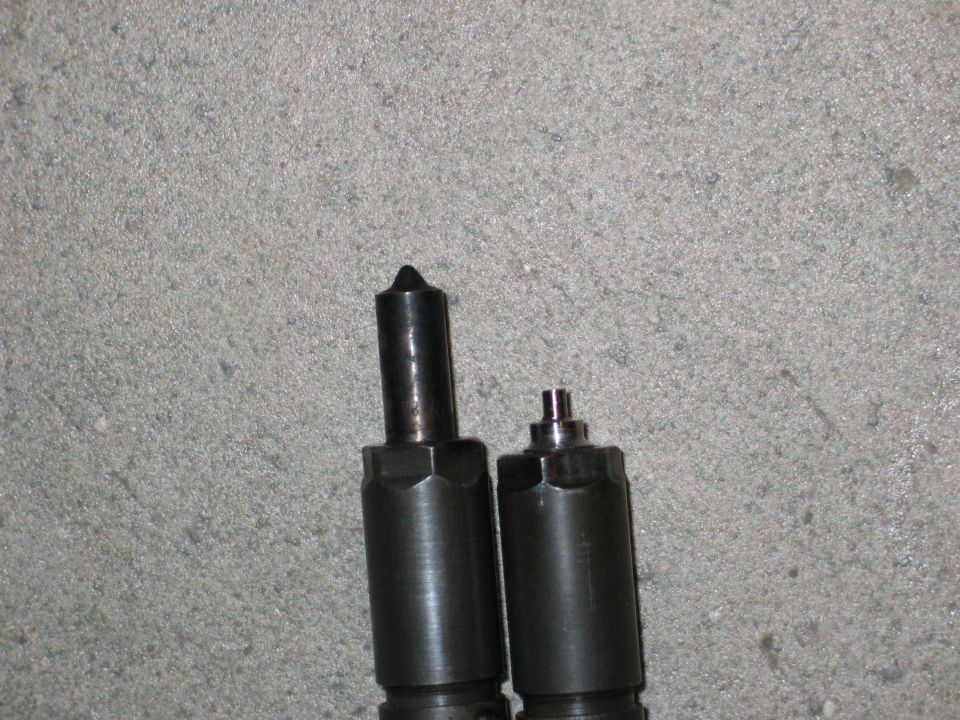 Two dark metal diesel injectors with hexagonal bases against a grey concrete background