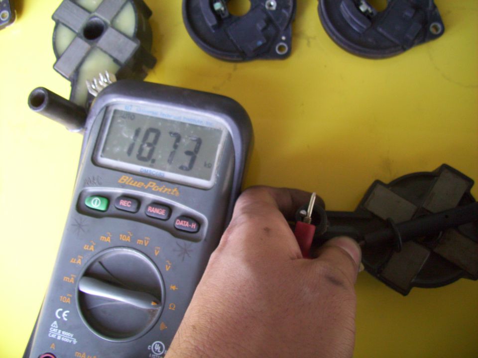 Hand holding multimeter probes to ignition coils, reading 18.73