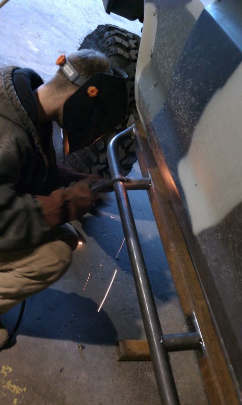 1979 Ford Bronco - person wearing a welding mask welds a tubular steel rock slider onto a vehicle's rocker panel, sparks visible.