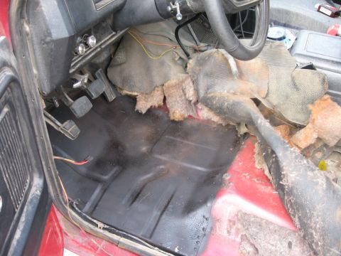 1981 Ford Bronco - The interior floor of a vehicle shows worn carpeting, exposed insulation, and the pedals and steering column.