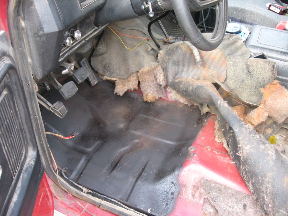 vehicle floor with torn carpet and exposed insulation, showing pedals and part of steering wheel