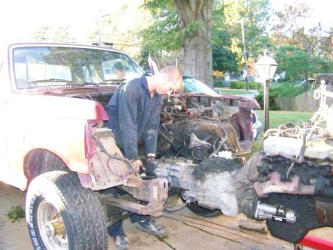 1985 Ford Bronco - Mechanic works on the disassembled front end of a red Ford Bronco, revealing the engine and transmission components.