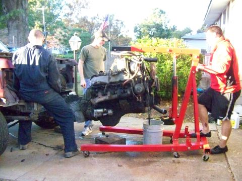 1985 Ford Bronco - Three men work together to remove an engine from a vehicle using a red engine hoist and a bucket.