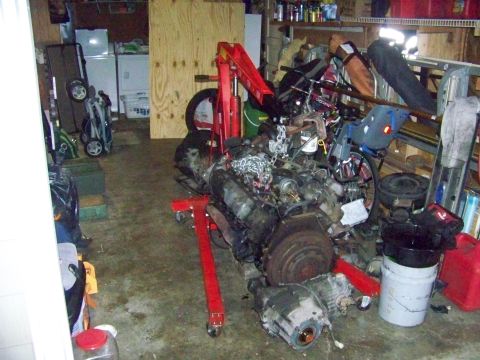 1985 Ford Bronco - An engine and transmission assembly is mounted on a red engine stand in a cluttered garage setting.