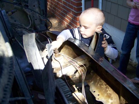 1985 Ford Bronco - A young child inspects the engine compartment of a vehicle, surrounded by wires and mechanical parts.