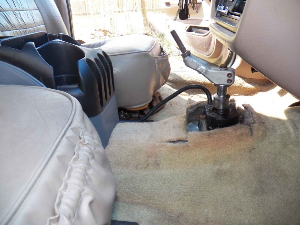 Aftermarket floor shifter installed through cut tan carpet between the seats of a tan-interior car
