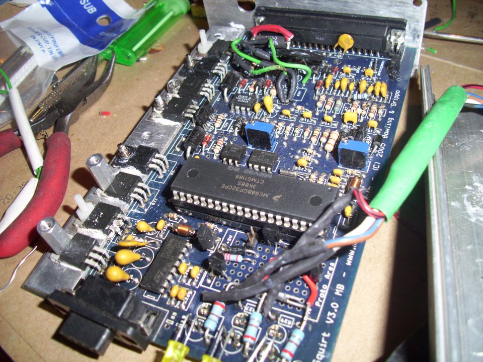 Close-up of a circuit board with a large black chip, resistors, capacitors, and wires. Tools and other electronic components are visible in the background