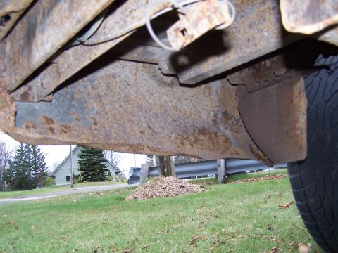 1969 Chevrolet C/K Pickup - Close-up view of a heavily rusted truck frame and a tire, with a house and trees in the background.