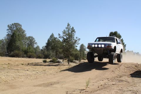 1996 Ford Bronco - This modified 1996 Ford Bronco is captured mid-jump during an off-road excursion, kicking up dust against a clear blue sky.