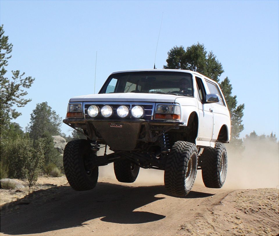 White 1996 Ford Bronco airborne on a dirt trail, modified with large tires and off-road bumper