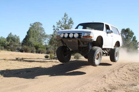 1996 Ford Bronco - A white first-generation-style full-size Ford Bronco SUV catches air mid-jump on a dusty dirt trail, showing its lifted suspension, large m…