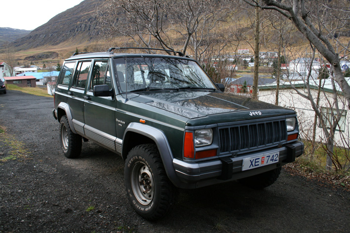 Green 1992 Jeep Cherokee parked on gravel road with houses and hills in background