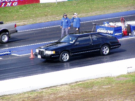 1999 Ford Crown Victoria - A black coupe is on the drag strip with two people standing nearby. Another vehicle is visible to the left.