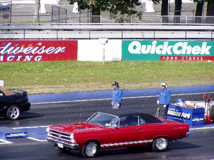 1999 Ford Crown Victoria - A red convertible sits on a drag strip with a black car to its left and two people standing nearby.
