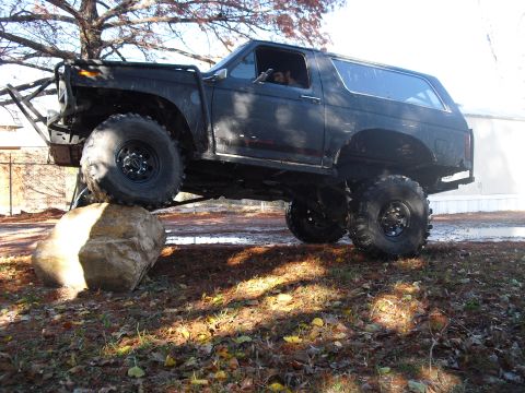1986 Ford Bronco - Modified black Ford Bronco with off-road tires and custom bumper is shown climbing over a large rock.