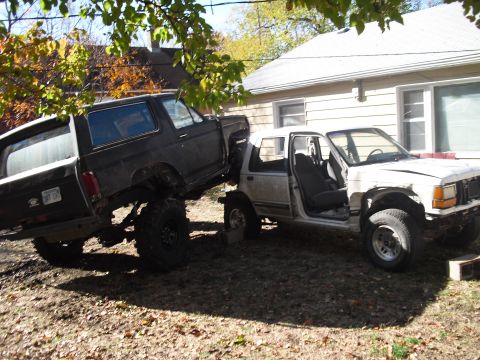 1986 Ford Bronco - Two SUVs are parked in a yard, one is lifted and the other is damaged.