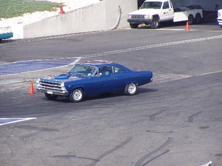 1999 Ford Crown Victoria - A blue 1966 Ford Fairlane coupe is on a race track, with a white truck and trailer in the background.