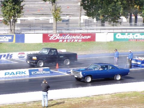 1999 Ford Crown Victoria - A blue classic car and a black pickup truck are lined up on a drag strip with spectators and advertising in the background.
