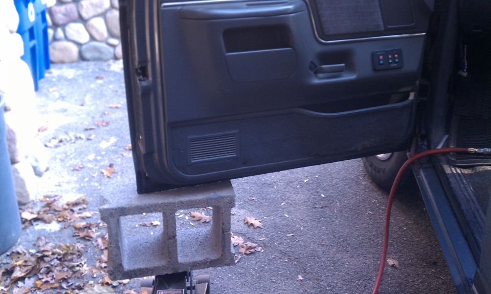 Open driver's door of dark Ford Bronco supported by cinder block on a floor jack in a driveway