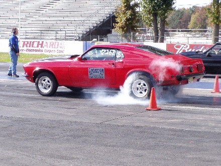 1999 Ford Crown Victoria - A red 1969 Ford Mustang performs a burnout at a drag strip, with a person watching and another car in the background.