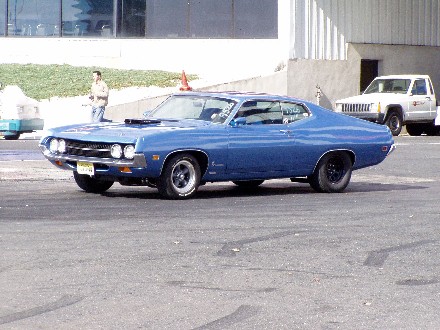 1999 Ford Crown Victoria - A blue 1970s muscle car is parked on asphalt, with a white Jeep visible in the background.
