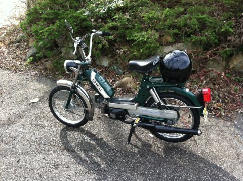 1981 Garelli Rally Sport - A green moped is parked on a gravel surface with bushes in the background. A black helmet rests on the seat.