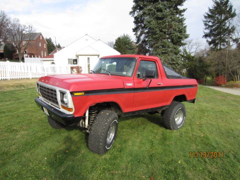 1979 Ford Bronco - Red full-size Ford Bronco with a black lower bodyside stripe and removed rear top, parked on a front lawn and photographed from a 3/4 front…