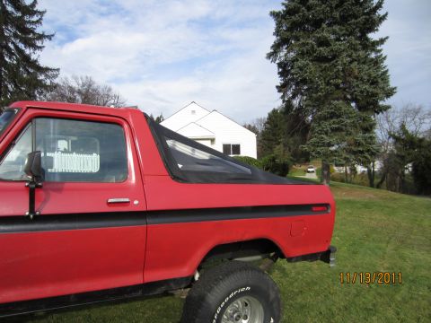 1979 Ford Bronco - A red SUV with a black soft top is parked on a grassy lawn with a house and trees in the background.
