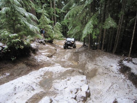 2008 Jeep Wrangler - A black Jeep Wrangler navigates a challenging off-road trail with mud and snow in a dense forest setting.