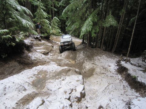 2008 Jeep Wrangler - A white off-road vehicle navigates a challenging, snow-dusted trail through a dense forest.